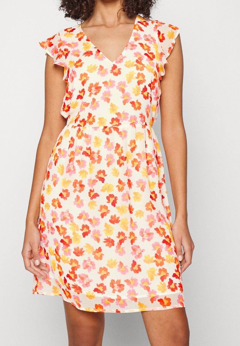 Donna che indossa un vestito senza maniche con scollatura a V, decorato con un motivo floreale rosso, arancione e rosa su sfondo bianco, in piedi con le mani rilassate.