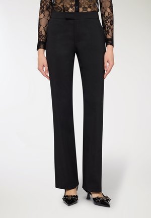 Pantaloni neri su misura con texture liscia e design a gamba dritta, abbinati a scarpe nere con punta a punta che presentano un fiocco e dettagli dorati.