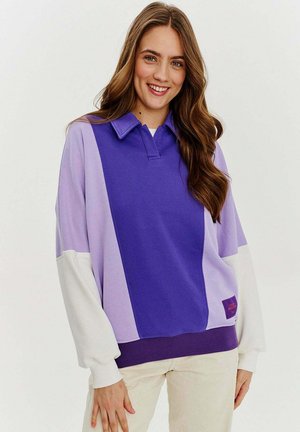 Svetrová mikina v purpurovej a levanduľovej farbe s golierom, širokými bielymi rukávmi a rebrovaným spodným okrajom. Na prednej strane sa nachádza logo.