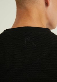 Czarna teksturowana dzianinowa koszulka z okrągłym dekoltem, z widocznym detalem szwów z tyłu. Tkanina wydaje się lekka i oddychająca.