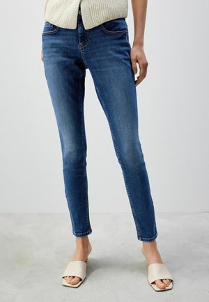 Wąskie niebieskie dżinsy ze stretchowego denimu z subtelnym blaknięciem, wyposażone w talię o średnim stanie, design z pięcioma kieszeniami, zestawione z beżowymi sandałami na płaskim obcasie.