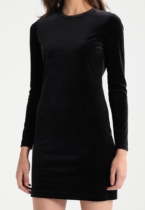 Mujer con un vestido negro de terciopelo ajustado de manga larga y cuello redondo, de pie frente a un fondo claro y liso.