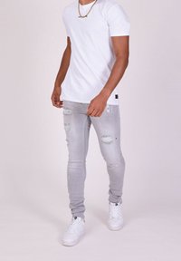 Camiseta de algodón blanca, de manga corta, combinada con unos vaqueros skinny gris claro desgastados que presentan rotos y zapatillas blancas. Collar de cadena dorada visible.