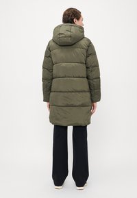 Personen som vender bort fra kameraet, iført en olivenfarget hettejakke med dunfyll, svarte bukser og hvite sko mot en enkel lys bakgrunn.