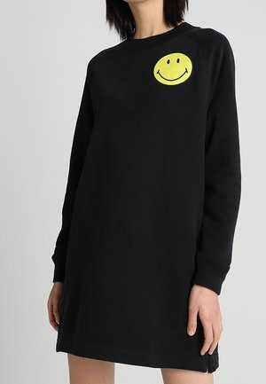 Zwarte mini-jurk met lange mouwen gedragen door een persoon, met een gele smiley-patch op de linker bovenkant van de borst.