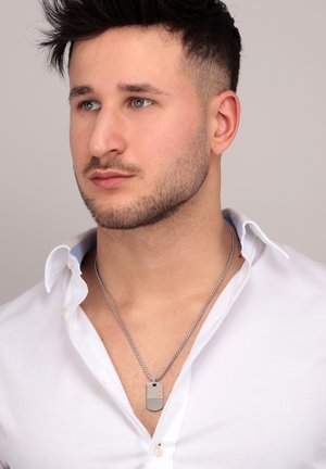 Collana con ciondolo dog tag in argento su una catena metallica, indossata sopra una camicia bianca. Il ciondolo presenta un design testurizzato e un piccolo accento dorato.