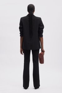 Černý přizpůsobený blazer s ohrnutými rukávy, v kombinaci s černými kalhotami na míru. Drží hnědou koženou psaníčko. Hladká textura a strukturovaný design.