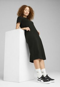 Abito nero fino al ginocchio con maniche corte, in tessuto testurizzato. Abbinato a sneakers bianche e nere e calzini bianchi a coste, appoggiato a un cubo bianco.
