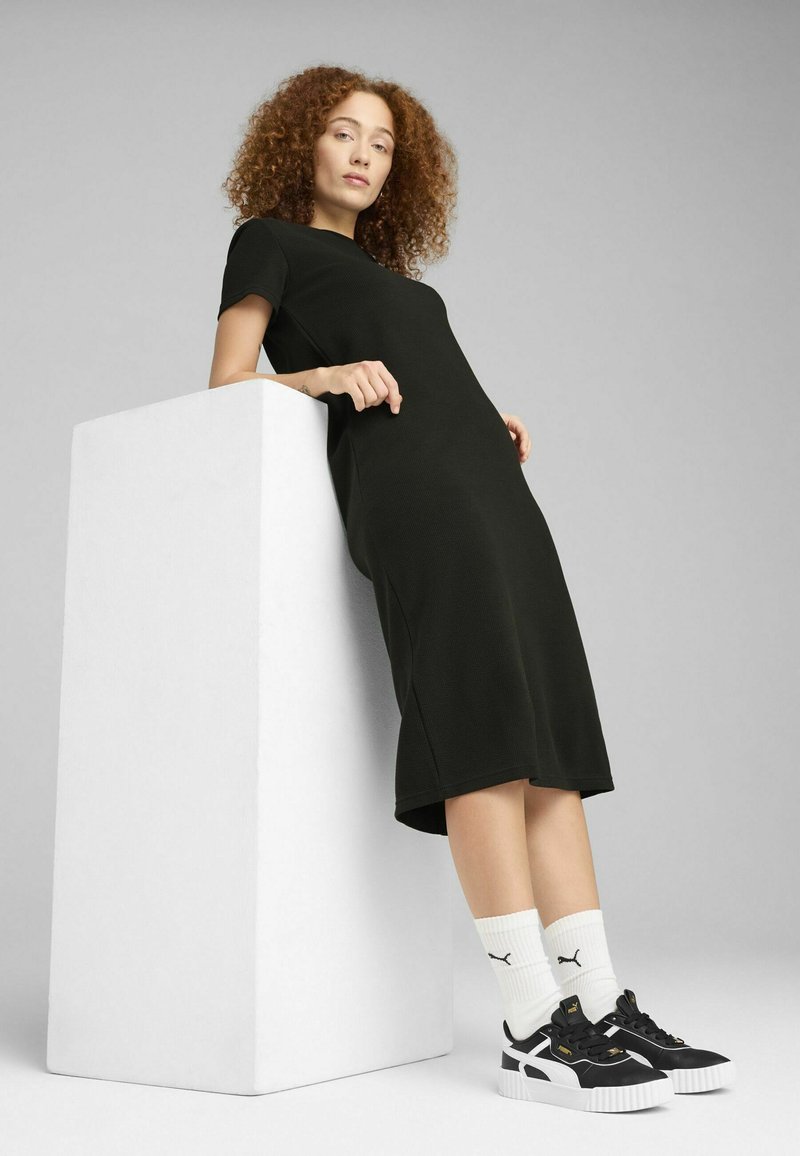 Robe noire mi-longue avec des manches courtes, en tissu texturé. Associée à des baskets noir et blanc et des chaussettes blanches côtelées, s'appuyant contre un cube blanc.