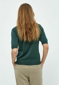 Suéter de manga corta verde oscuro con un acabado texturizado y un diseño ajustado, combinado con pantalones beige claros que cuentan con bolsillos traseros.