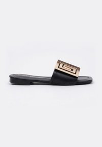 Sandalia plana negra con una cinta ancha y un acento de hardware dorado rectangular en la parte superior. Textura de cuero suave con poco grabado.