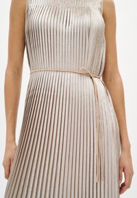 Beżowa plisowana sukienka wykonana z błyszczącego materiału, z dopasowanym górnym partem i paskiem wiązanym w talii. Tekstura jest gładka z pionowymi fałdami.