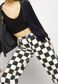 Un débardeur en maille côtelée noir coupé sous un sweat à capuche zippé sombre, associé à un pantalon à carreaux noir et blanc à taille haute et coupe loose.