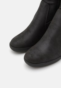 Bottines noires en matériau synthétique lisse, bout rond, texture subtile, et semelle en caoutchouc basse et flexible. Équipées de panneaux élastiques latéraux.