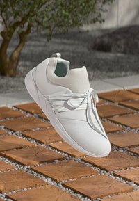 Chaussure de sport blanche avec une tige lisse, texture en maille et lacets élastiques. Dispose d'un talon rembourré et d'un design minimaliste.