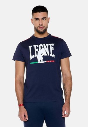Homme aux cheveux courts et foncés avec barbe, portant un t-shirt bleu marine Leone avec un logo de boxe et les couleurs du drapeau italien, debout devant un fond uni.