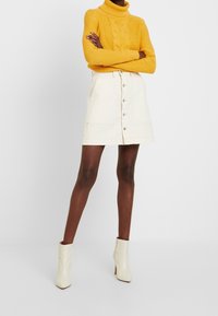 Gestrickter gelber Rollkragenpullover kombiniert mit einem cremefarbenen Denimrock mit Knöpfen vorne, Seitentaschen und geradem Schnitt. Weiße Stiefeletten.