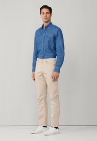 Lichtblauwe denim overhemd met knoopsluiting en zak; beige broek; witte sneakers; model poseert tegen een neutrale achtergrond.