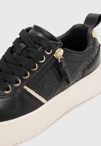Basket noire avec une tige en cuir texturé, accents en métal doré, semelle blanche et fermeture lacée avec une fermeture éclair latérale supplémentaire.