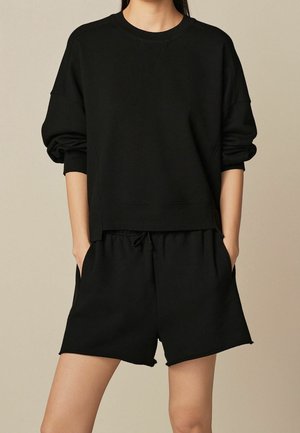 Personne portant un sweat-shirt noir à manches longues et un short noir avec cordon, debout les mains dans les poches devant un fond beige.