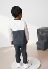 Il bambino indossa una felpa a blocchi di colore con sezioni grigie e bianche, abbinata a pantaloni scuri con strisce laterali bianche e sneaker bianche.