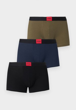 Modèle masculin debout pieds nus sur un fond uni, portant un boxer vert olive avec une ceinture noire et une étiquette logo rouge.