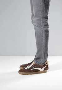 Braune Wildleder-Sneaker mit weißen Akzenten und strukturierten Gummisohlen. Verfügen über Schnürsenkel und ein niedrig geschnittenes Design. Getragen mit grauen Slim-Fit-Jeans.
