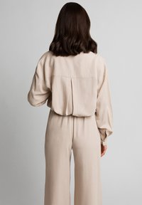 Beżowa bluzka z długim rękawem o luźnym kroju, z plisowanym detalem z tyłu i guziczkami przy mankietach, w zestawie ze spodniami o szerokich nogawkach w tym samym kolorze. Gładka tekstura.