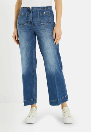 Mujer con vaqueros azules de tiro alto y pernera recta, con bolsillos delanteros y zapatillas blancas, posando frente a un fondo liso.