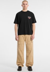 T-shirt nera in cotone con logo rosso e bianco, abbinata a pantaloni cargo beige dotati di molteplici tasche e vestibilità ampia. Scarpe nere completano il look.