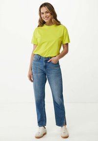 Limetkově zelené tričko s krátkým rukávem v kombinaci se světle modrými džíny rovného střihu s bočními rozparky, nošené s bílými teniskami na jednoduchém pozadí.