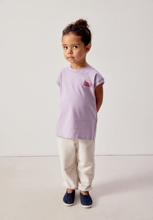 Mladé dítě s kudrnatými vlasy sepnutými do drdolu, oblečené v lila tílku s nápisem "American Vintage", bílých kalhotách a tmavě modrých sliponech, stojící.