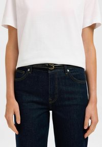 Mustavyö, jossa on kultapala, yhdistetty valkoiseen lyhythihaiseen t-paitaan ja tummansinisiin farkkuihin, joissa on erottuva keltainen tikkaus.