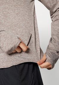 Dugim rukavima, smešani smešani smeđi top s otvorima za palčeve, složen preko crnih sportskih hlača. Teksturirana tkanina s detaljem bočne džepa.