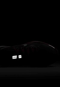 Chaussure de sport noire avec des accents rouges, présentant une surface texturée, des lacets et un logo Nike réfléchissant sur le côté, affichée sur un fond sombre.