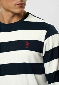 Gestreepte langlijf shirt in marineblauw en witte katoen met een geribbelde ronde hals. Heeft een rode geborduurde logo op de linkerborst.