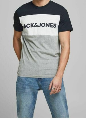 Homme portant un t-shirt Jack & Jones noir, blanc et gris assorti à un jean bleu, debout devant un fond gris uni.