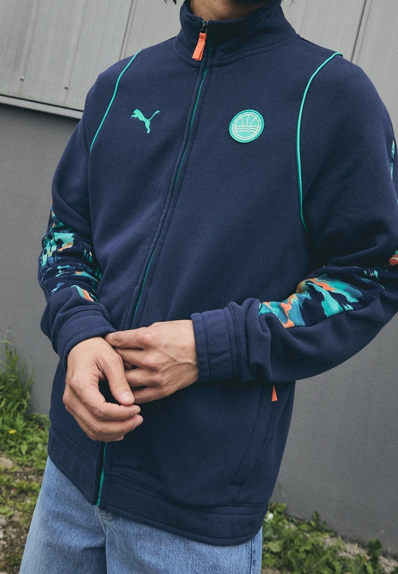 Hombre vistiendo una chaqueta con cremallera de color azul marino con acentos turquesa y naranja, de pie al aire libre con las manos juntas al frente, frente a una pared gris.