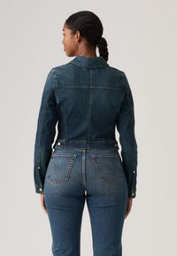 Γυναίκα με σκούρα πλεξούδα, φοράει εφαρμοστή σκούρα μπλε τζιν ζακέτα και ταιριαστό τζιν παντελόνι, φαίνεται από πίσω με φόντο μια απλή επιφάνεια.