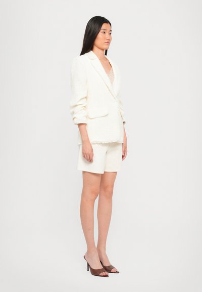 Witte tweed blazer met rafelige randen, gestructureerde schouders en twee voorzakken, gecombineerd met crèmekleurige shorts en bruine open teen haksandalen.