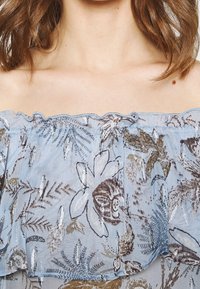 Haut bleu clair à épaules dénudées en tissu transparent. Présente des motifs floraux en brun et or avec un col à volants et une légère brillance.