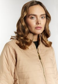 Tan gewatteerde pufferjas met een hoge kraag, gouden rits en subtiele glans. Gladde textuur, zichtbare stiksels en een getailleerd ontwerp.