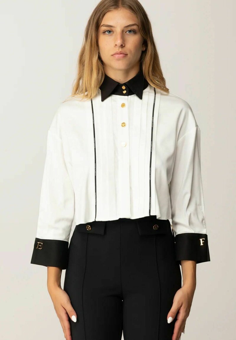 Camicia bianca corta con colletto e polsini neri, caratterizzata da bottoni dorati e dettagli plissettati. Pantaloni neri a vita alta con dettagli dorati.