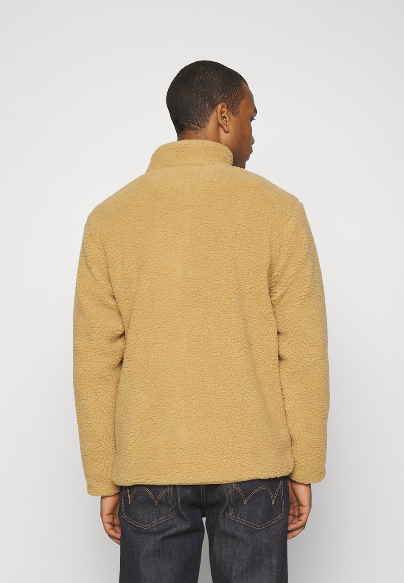 Giacca in pile color sabbia con colletto alto, superficie texturizzata e maniche lunghe. Indossata con jeans in denim scuri, che mostrano un taglio rilassato.