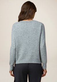 Femme aux longs cheveux bruns portant un pull à manches longues gris clair avec de petits clous métalliques sur l'encolure et les manches, assorti à un pantalon foncé.