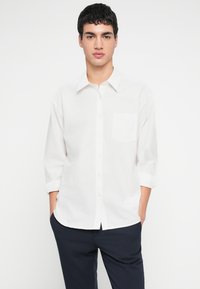 Fehér gombos ing egyetlen mellzsebbel, hosszú ujjakkal, amiket könyöknél felhajtottak, könnyű anyagból készült. Sötét nadrággal viselve.