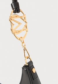 Black leather bag detail featuring a gold-tone metal logo hardware and a clasp. The finish is smooth with stitching accents.