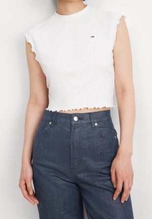 Biały, bezrękawnik z falbankami na krawędziach i małym logo flagi, w połączeniu z wysokim stanem ciemnoniebieskich dżinsów na stojącej osobie.