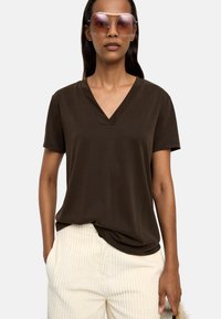 Brązowa koszulka typu v-neck z krótkim rękawem, zestawiona z jasnymi, prążkowanymi kremowymi spodniami. Modelka nosi oversize'owe, kolorowe okulary przeciwsłoneczne.