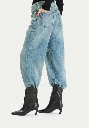 Persona che indossa jeans di denim azzurro chiaro larghi infilati negli stivali in pelle neri con tacco fino al polpaccio, con una mano nella tasca posteriore.